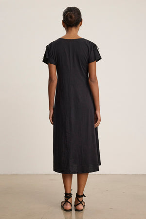 A woman faces away, wearing the VERONICA DRESS by Velvet by Graham & Spencer—a black, short-sleeve, loose-fit t-shirt dress—paired with black lace-up sandals, set against a plain light background.