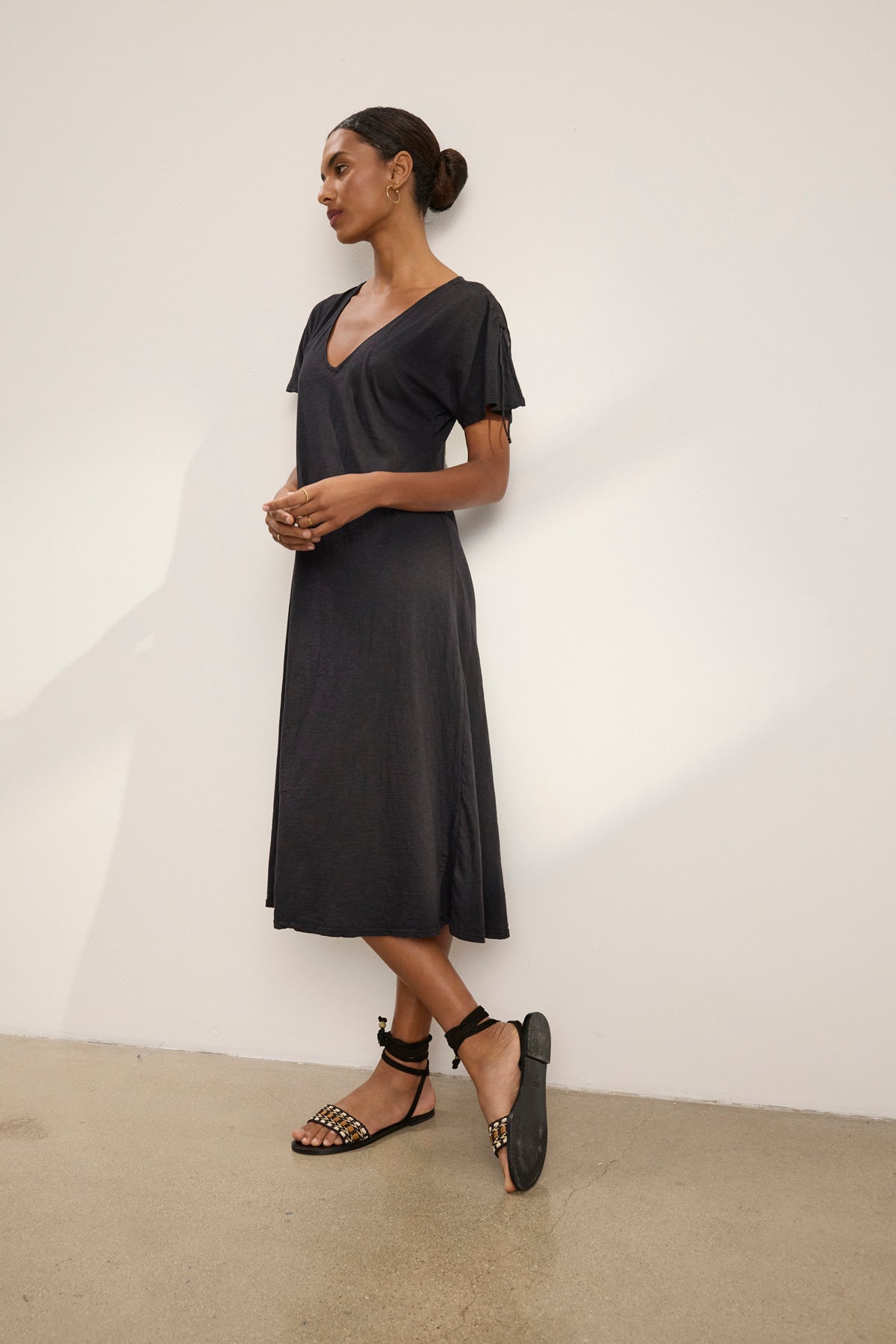 A woman stands against a plain wall, wearing the VERONICA DRESS by Velvet by Graham & Spencer— a versatile short-sleeve black midi t-shirt dress—paired with black strappy flat sandals, her hair styled in a neat bun.-39148782190785