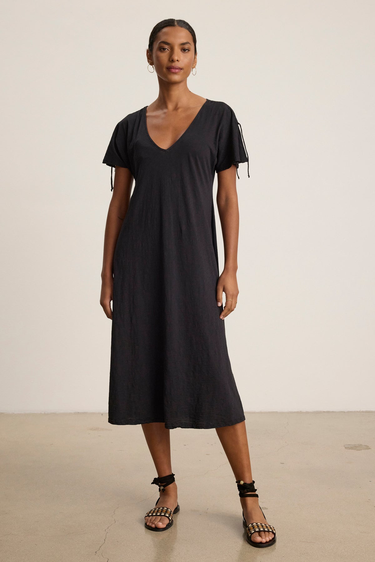 A woman stands indoors wearing the Velvet by Graham & Spencer VERONICA DRESS, a loose-fitting, short-sleeve black t-shirt dress with a v-neckline and tie details, paired with black flat sandals.-39148782158017
