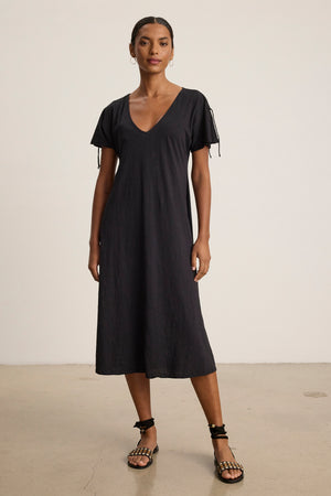 A woman stands indoors wearing the Velvet by Graham & Spencer VERONICA DRESS, a loose-fitting, short-sleeve black t-shirt dress with a v-neckline and tie details, paired with black flat sandals.
