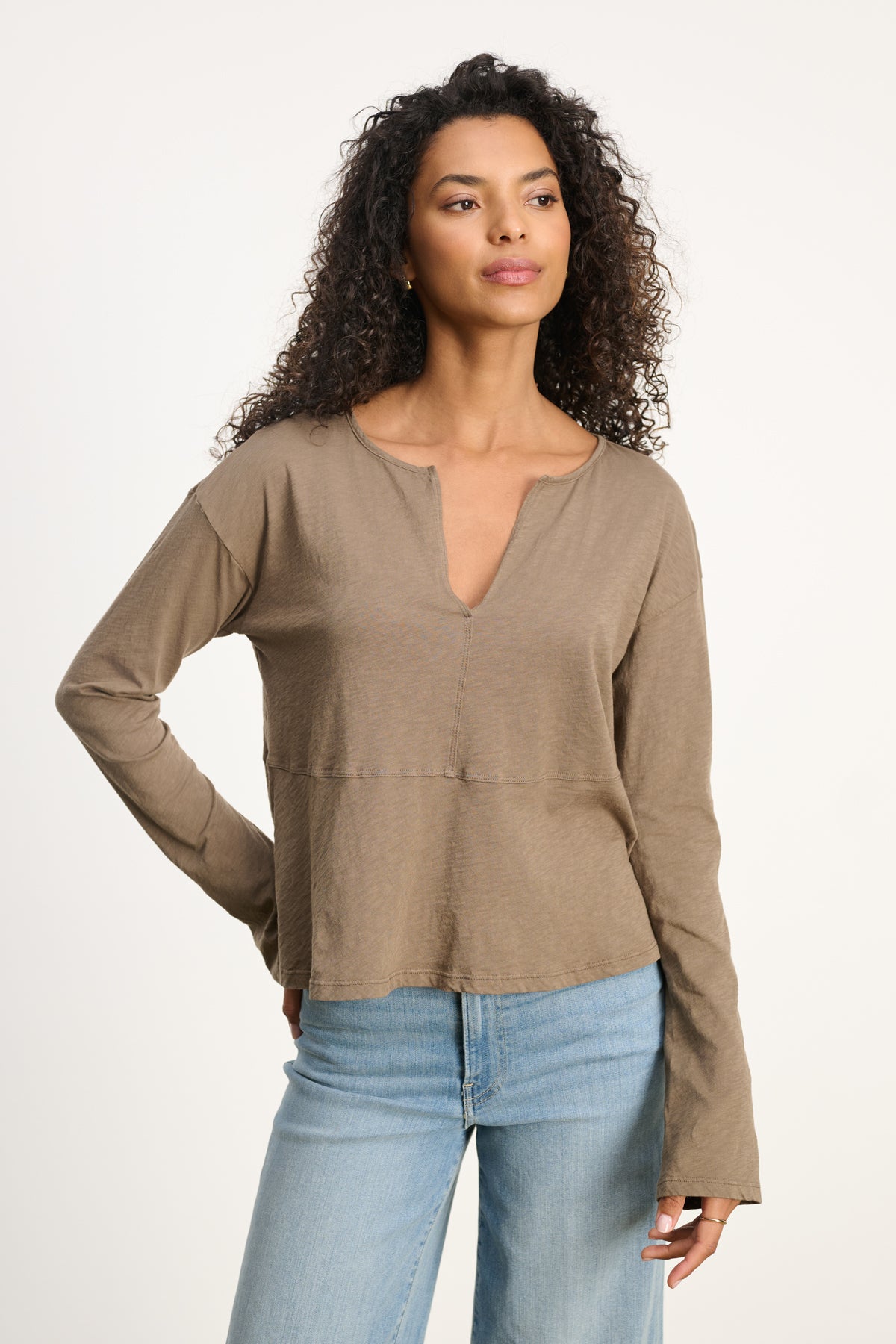 A woman with curly hair wears the VINT TEE by Velvet by Graham & Spencer—a relaxed fit, light brown cotton slub long sleeve with a notched neckline—paired with light blue jeans, posed against a plain backdrop.-39800429641921