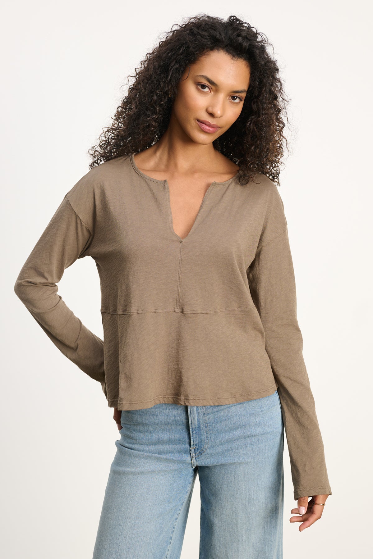 A woman with curly hair wears the VINT TEE by Velvet by Graham & Spencer, a relaxed fit long sleeve in vintage cotton slub, paired with light blue jeans against a plain light background.-39800429609153
