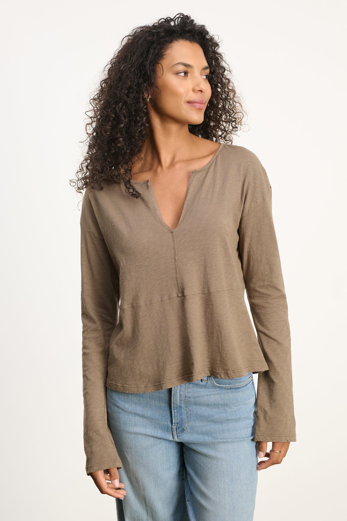A woman with curly hair models the VINT TEE by Velvet by Graham & Spencer in taupe—a relaxed-fit, long sleeve top—paired with light blue jeans, standing slightly to the side against a plain background.