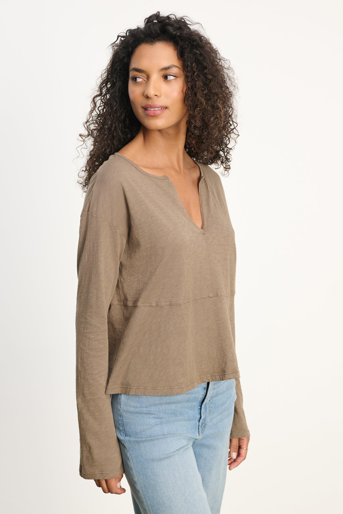 A woman with curly hair wears the Velvet by Graham & Spencer VINT TEE, a relaxed-fit vintage cotton slub long sleeve top in brown, paired with light blue jeans, against a plain light background and looking to the side.-39800429543617