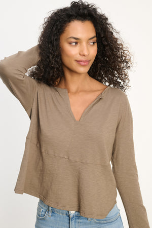 A woman with curly hair wears the Velvet by Graham & Spencer VINT TEE, a relaxed fit, long sleeve taupe top, paired with blue jeans. She stands with one hand behind her head, looking to the side.