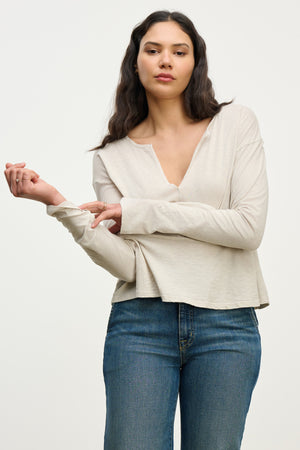 A woman poses against a plain background in the VINT TEE by Velvet by Graham & Spencer, a relaxed fit vintage cotton slub long sleeve beige top, paired with blue jeans and holding one sleeve with her arm bent.