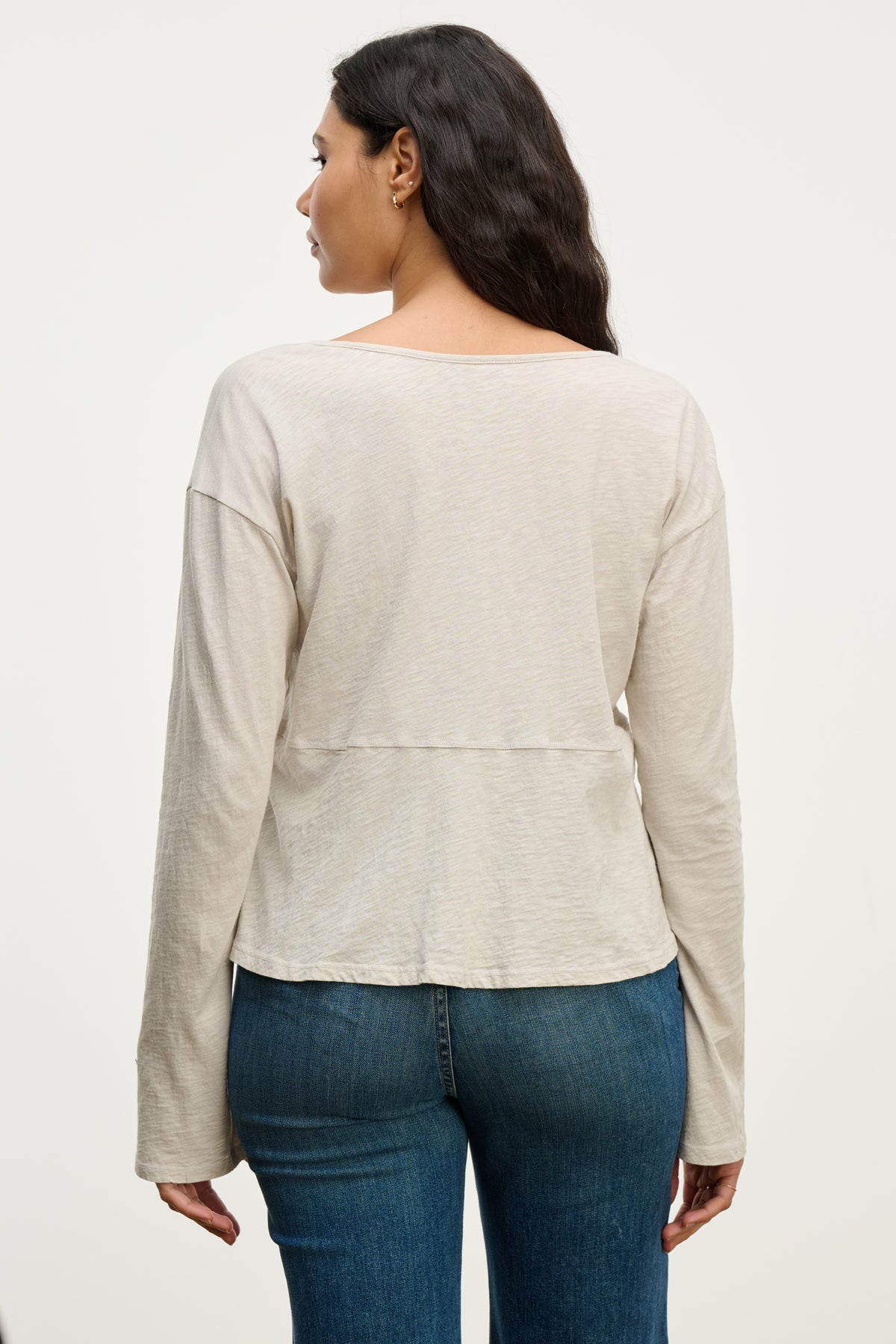   A woman stands facing away, wearing the VINT TEE by Velvet by Graham & Spencer—a relaxed fit long sleeve beige top—with blue jeans against a plain light background. 
