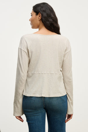 A woman stands facing away, wearing the VINT TEE by Velvet by Graham & Spencer—a relaxed fit long sleeve beige top—with blue jeans against a plain light background.