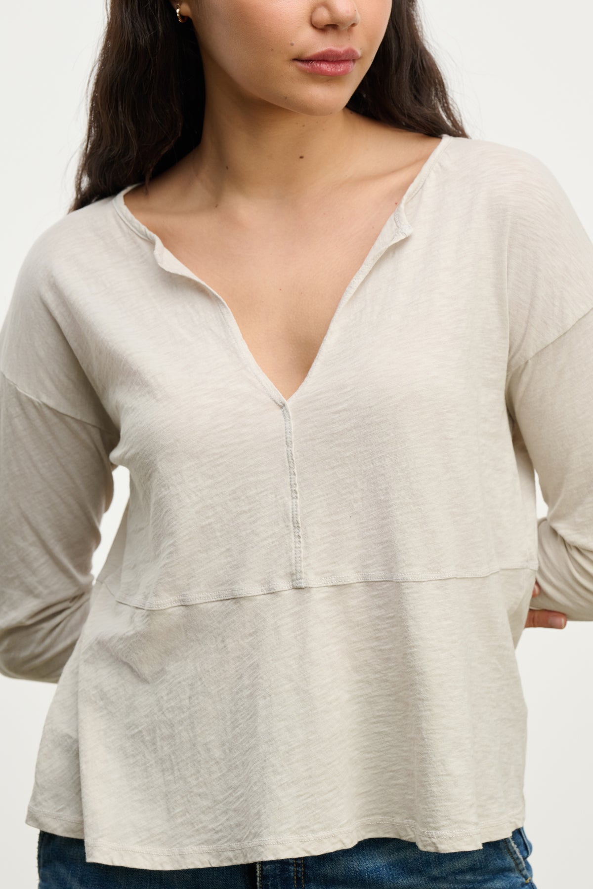   A woman wears the Velvet by Graham & Spencer VINT TEE, a vintage cotton slub long sleeve with a relaxed fit, paired with blue jeans, standing against a plain background with one hand behind her back. 