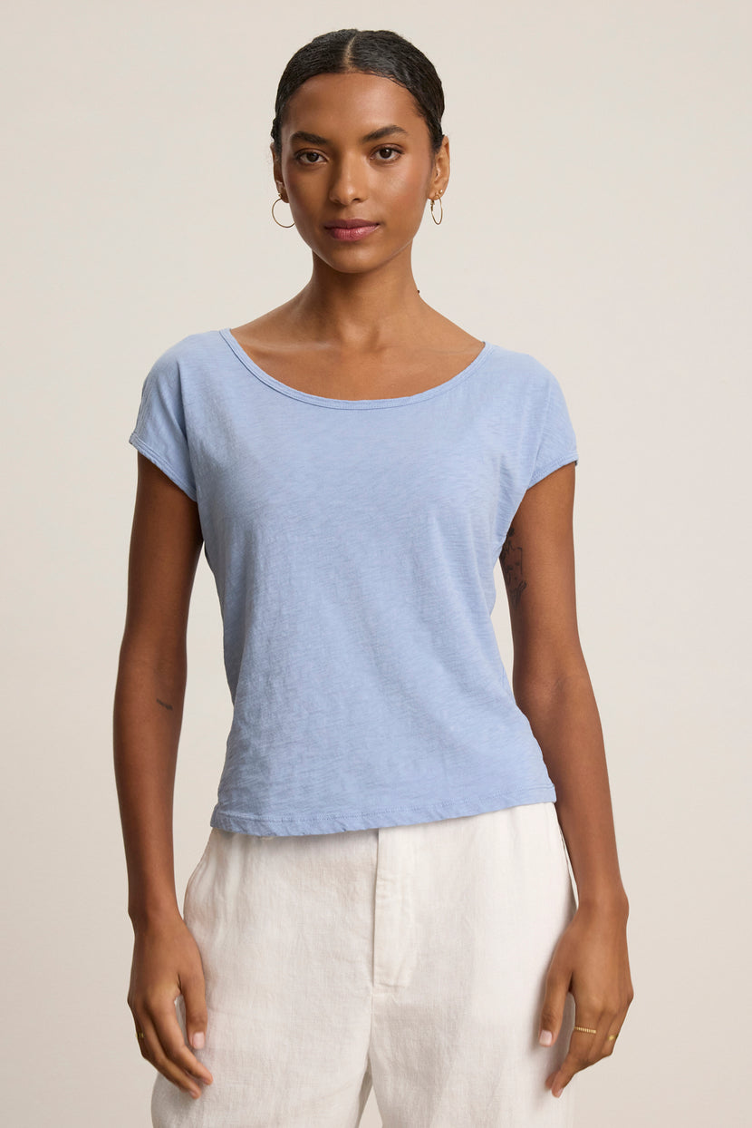 A woman stands against a neutral background, wearing the VIVIENNE TEE by Velvet by Graham & Spencer—a light blue cotton slub tee with a scoop neckline—paired with white pants, facing forward with a neutral expression.