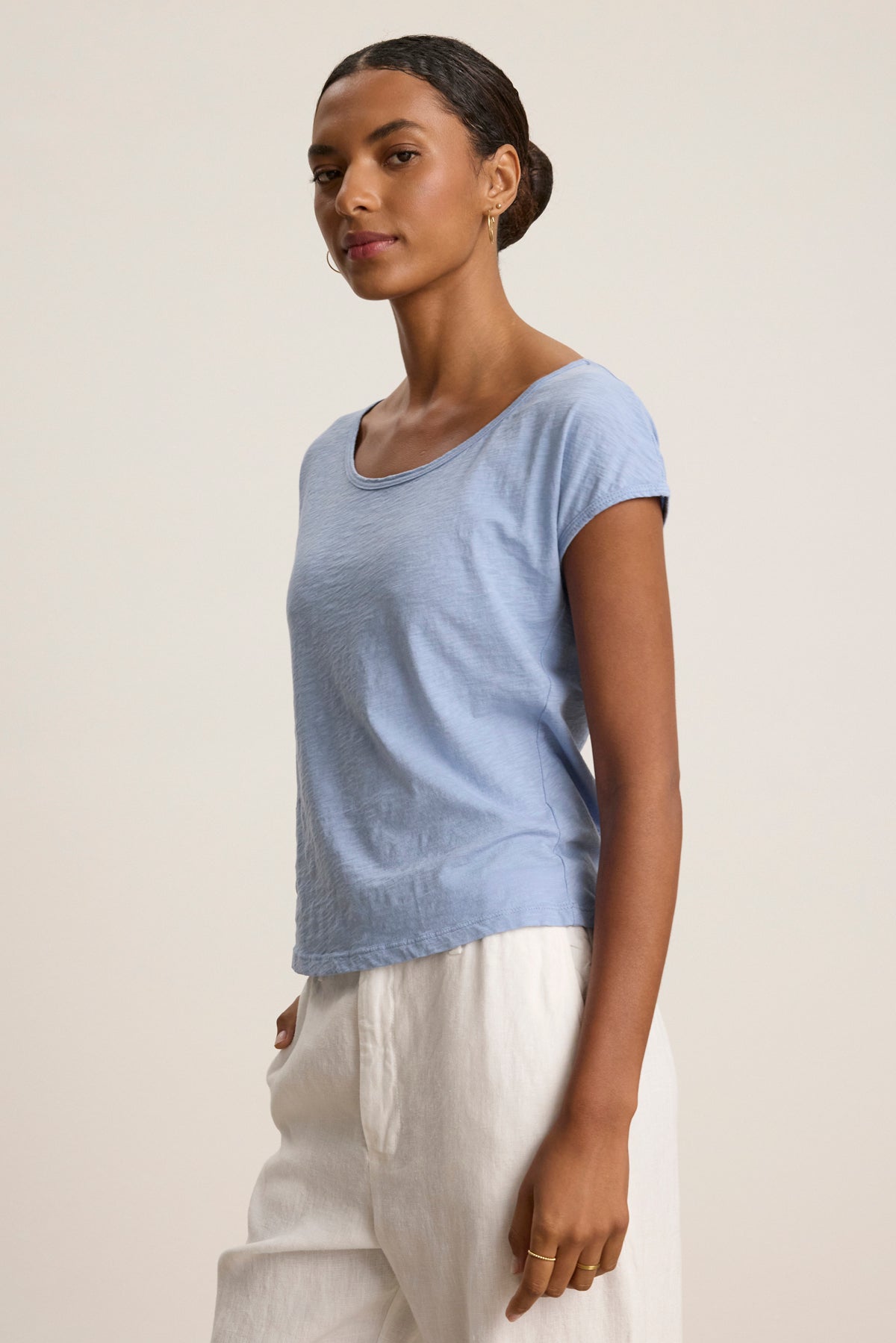 A woman poses in a relaxed stance, wearing the VIVIENNE TEE by Velvet by Graham & Spencer with a scoop neckline and white pants, set against a plain beige background.-39080058945729