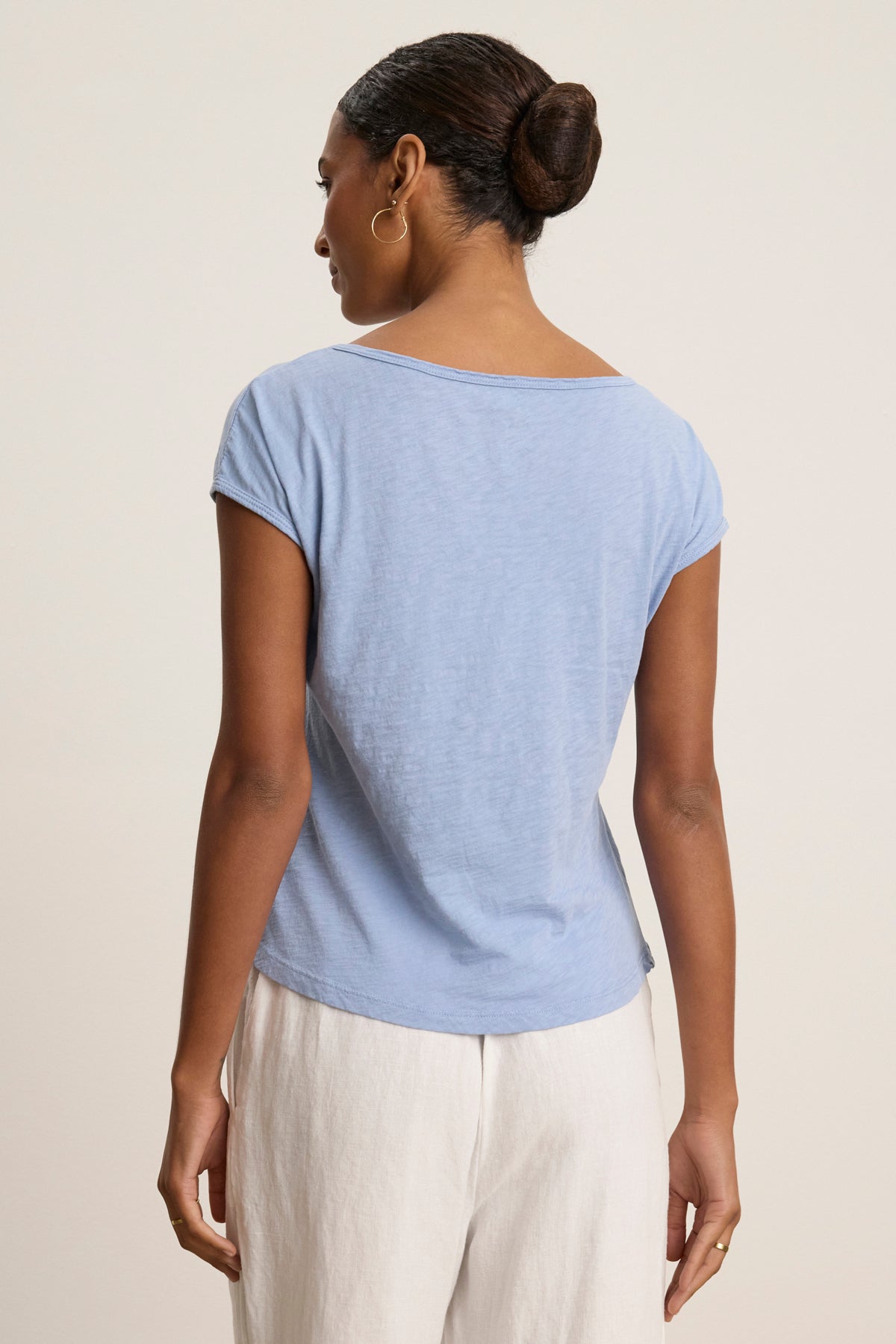 A woman faces away, wearing the VIVIENNE TEE by Velvet by Graham & Spencer in light blue cotton slub with a scoop neckline, paired with white pants. Her hair is in a bun against a plain background.-39080059011265