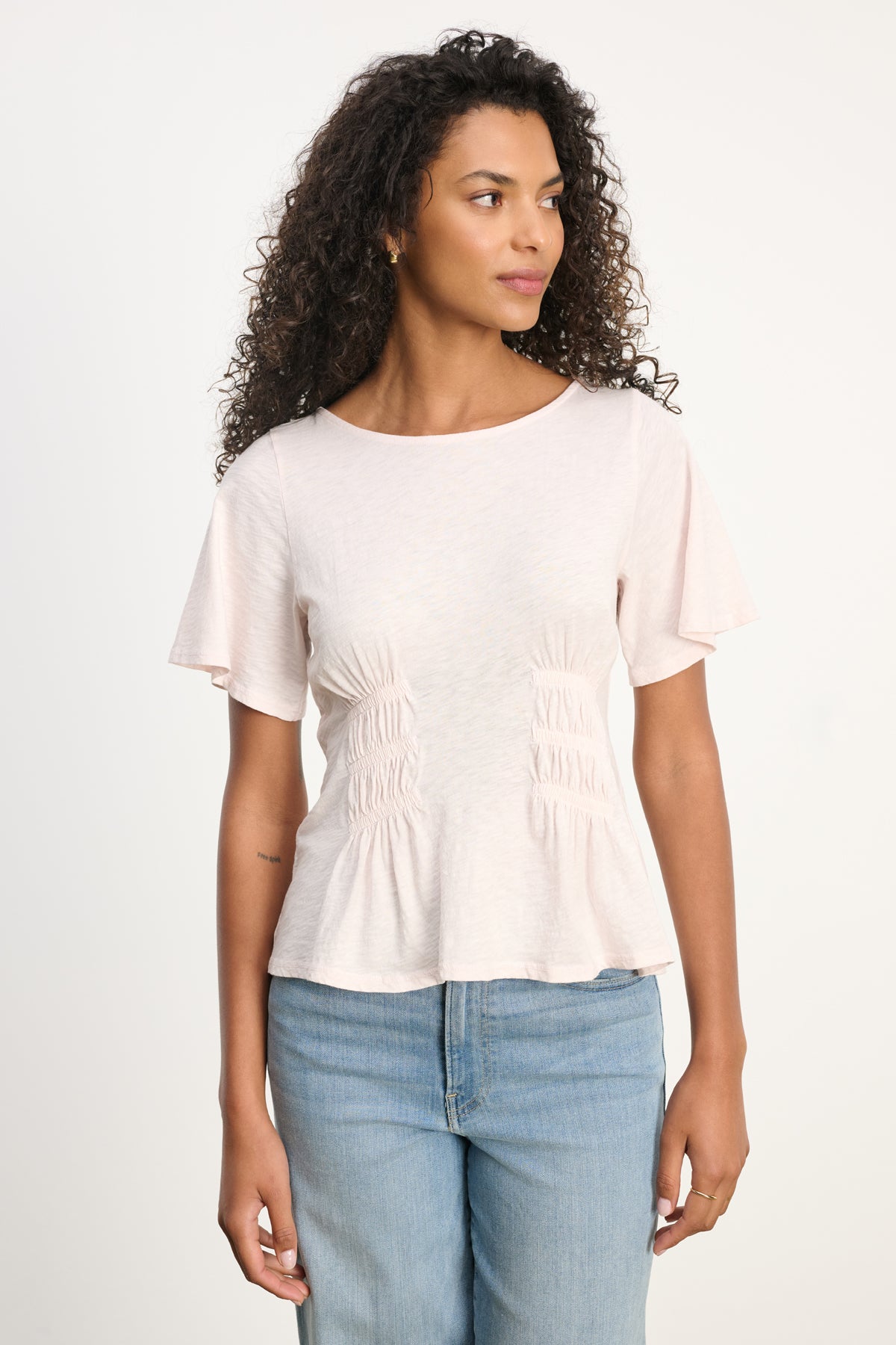 A woman with curly hair wears the Velvet by Graham & Spencer YASMINE TEE in light pink with blue jeans, standing against a plain light background and looking slightly to her left.-39617832386753