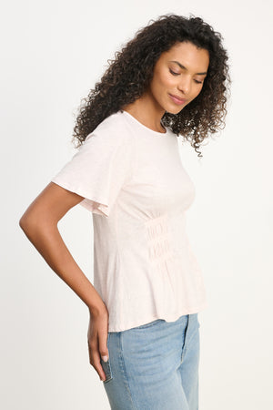 A woman with curly hair stands sideways against a white background, wearing the Velvet by Graham & Spencer YASMINE TEE in light pink cotton slub and blue smocked-waist jeans.