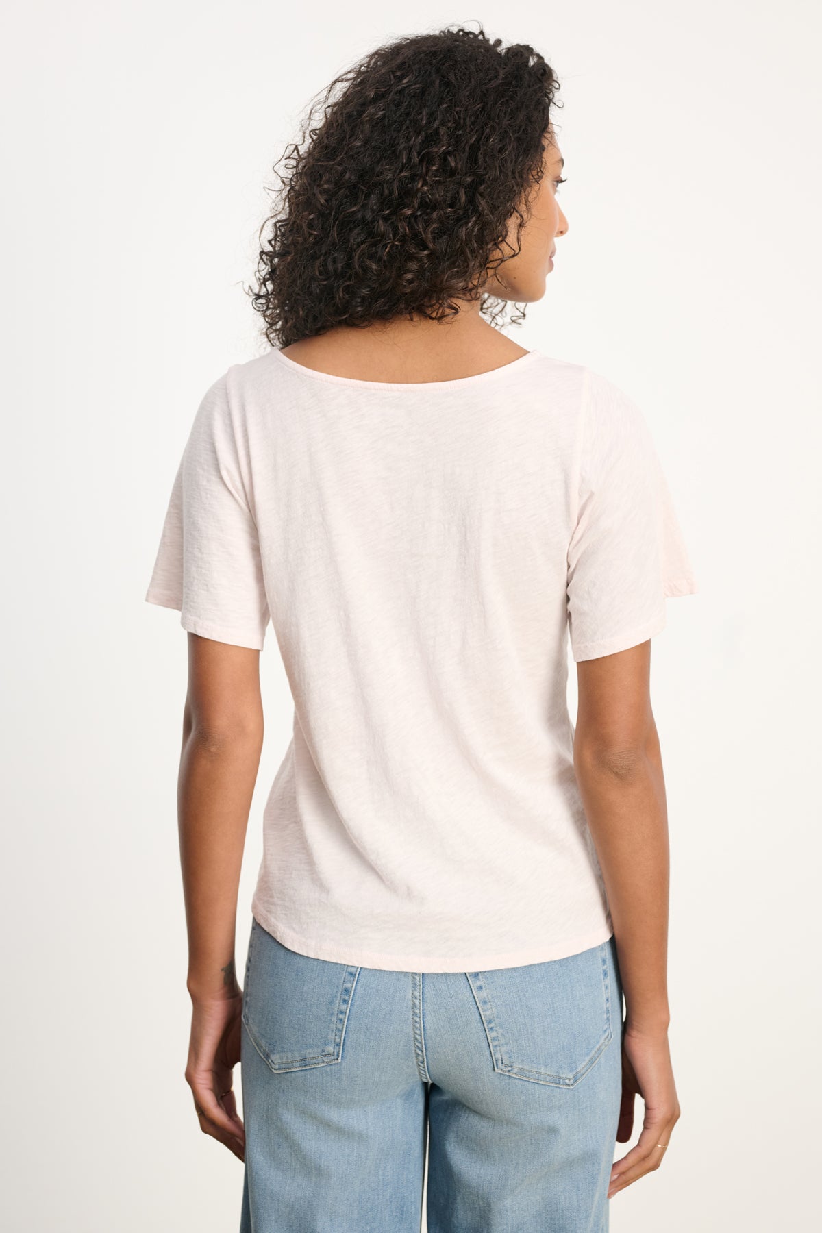 A person with curly hair is seen from the back wearing the YASMINE TEE by Velvet by Graham & Spencer in light pink, paired with light blue jeans, set against a plain background.-39617832288449