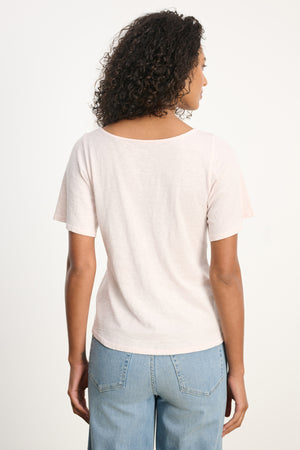 A person with curly hair is seen from the back wearing the YASMINE TEE by Velvet by Graham & Spencer in light pink, paired with light blue jeans, set against a plain background.