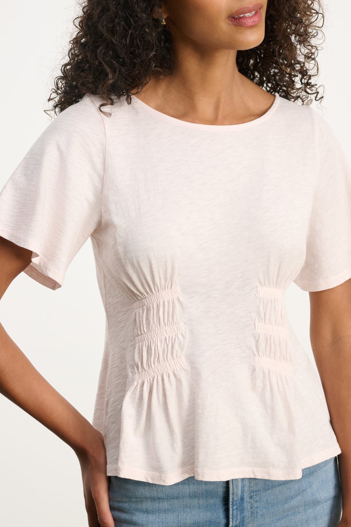 A woman wears the YASMINE TEE by Velvet by Graham & Spencer—a light pink, lightweight top featuring front ruching and a smocked elastic waist—paired with light blue jeans.-39617832255681