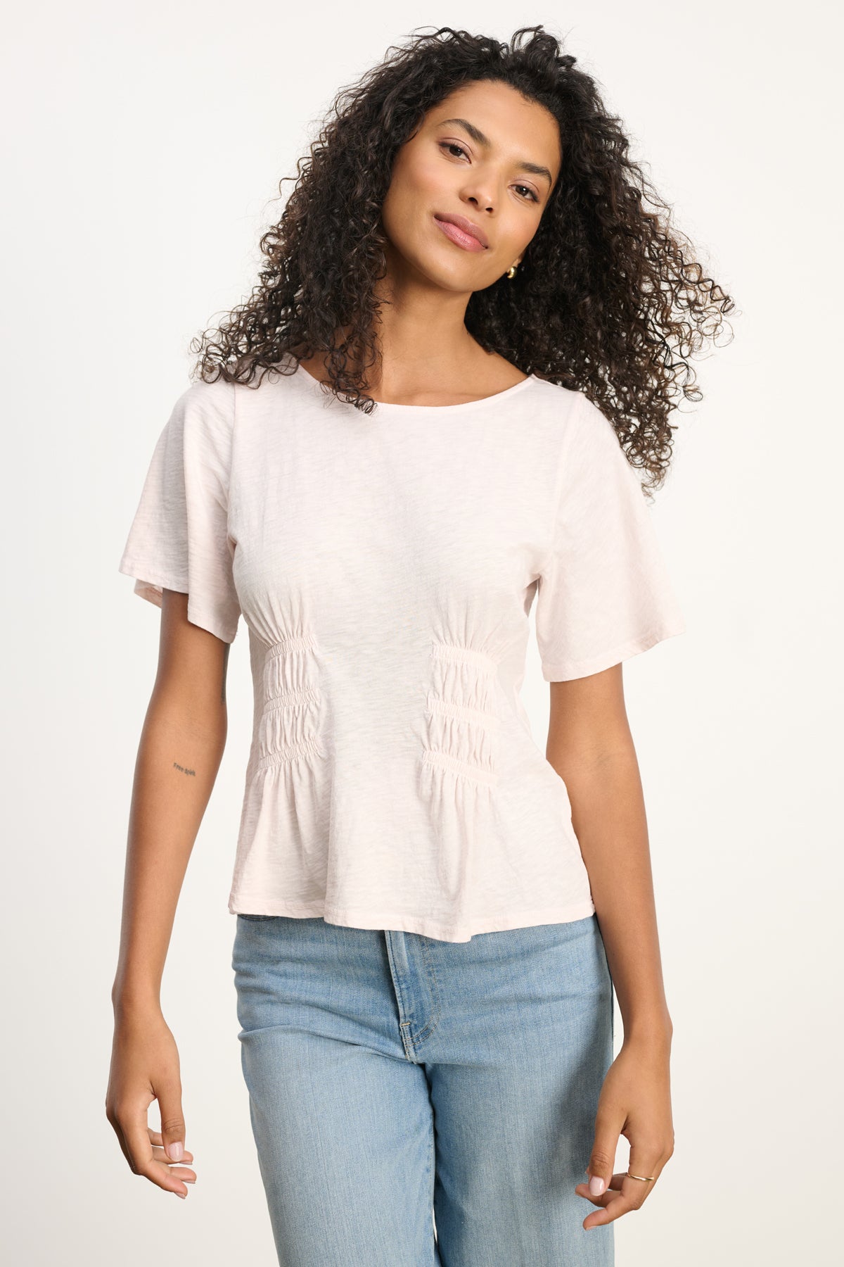 A woman with curly hair wears the YASMINE TEE by Velvet by Graham & Spencer, a vintage cotton slub light pink short-sleeve tee, paired with light blue jeans with a smocked elastic waist, standing against a plain background.-39617832157377