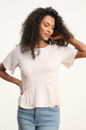 A woman with curly hair, wearing the Velvet by Graham & Spencer YASMINE TEE in light pink and blue jeans, stands against a plain background, touching her hair.