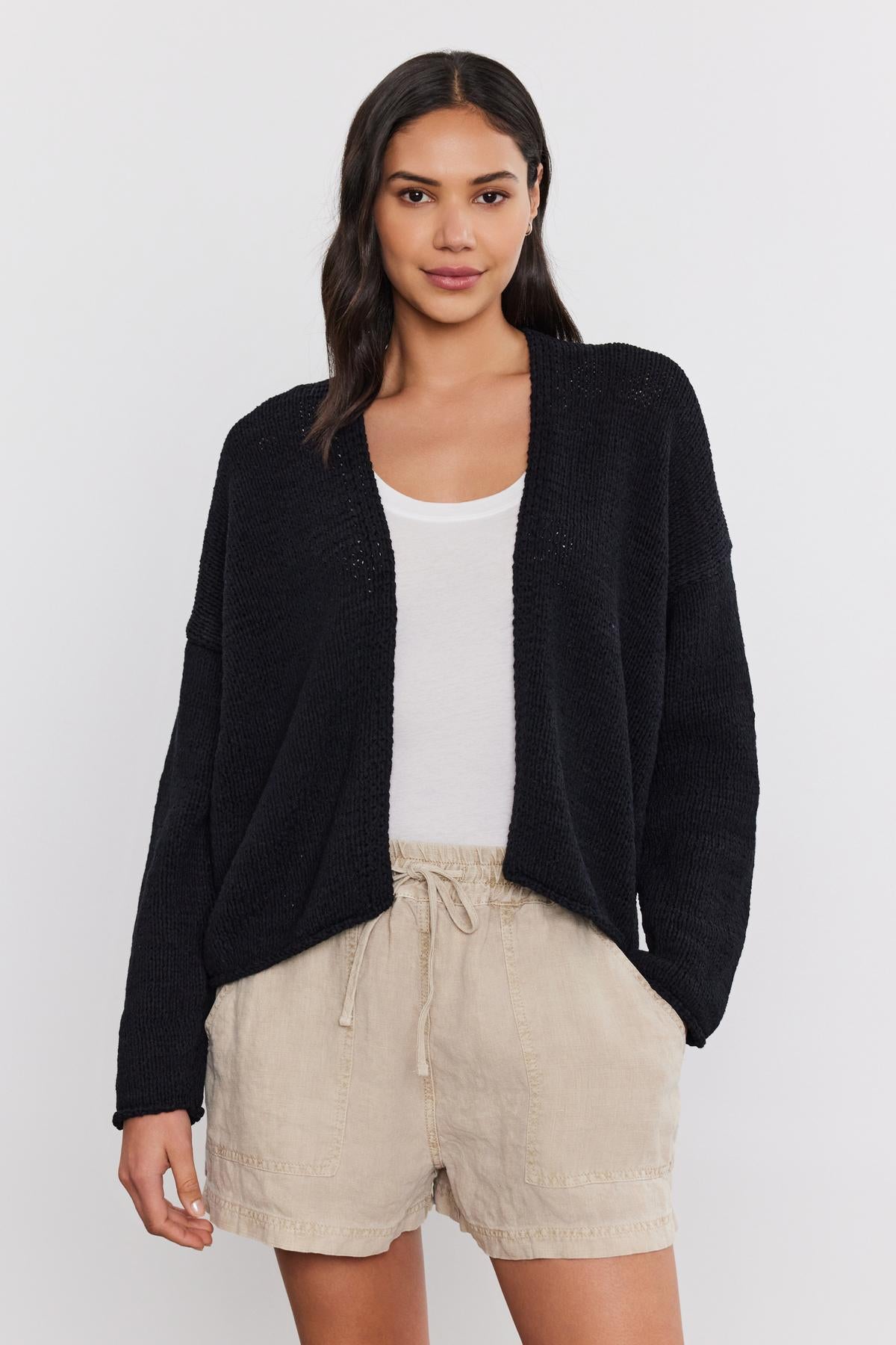 A woman wearing a black Velvet by Graham & Spencer HOLLIE cardigan, white top, and beige shorts, standing against a plain background.-36910017413313