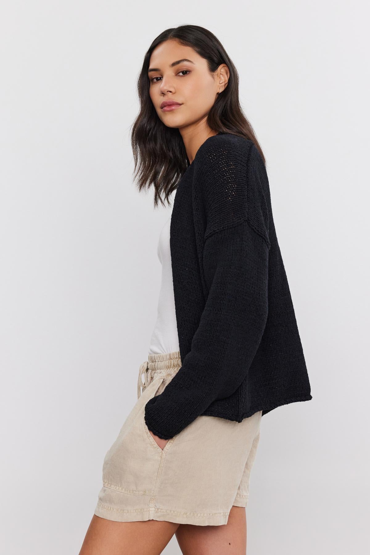 A woman in a casual outfit, featuring a Velvet by Graham & Spencer HOLLIE CARDIGAN over a white top and beige shorts, glancing to the side.-36910017446081