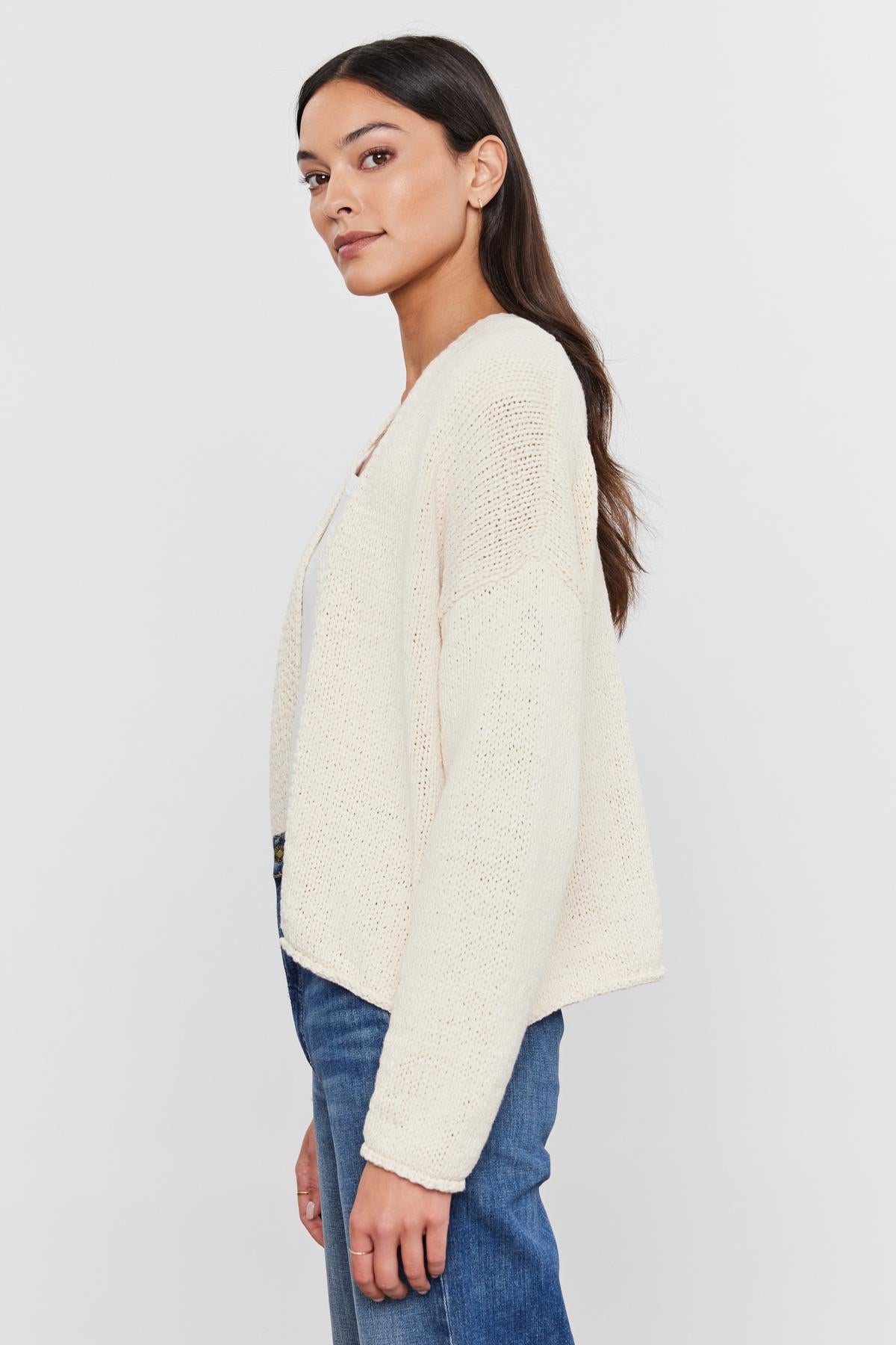 Woman in a "Velvet by Graham & Spencer" HOLLIE CARDIGAN and blue jeans standing sideways, looking at the camera over her shoulder.-36752930472129