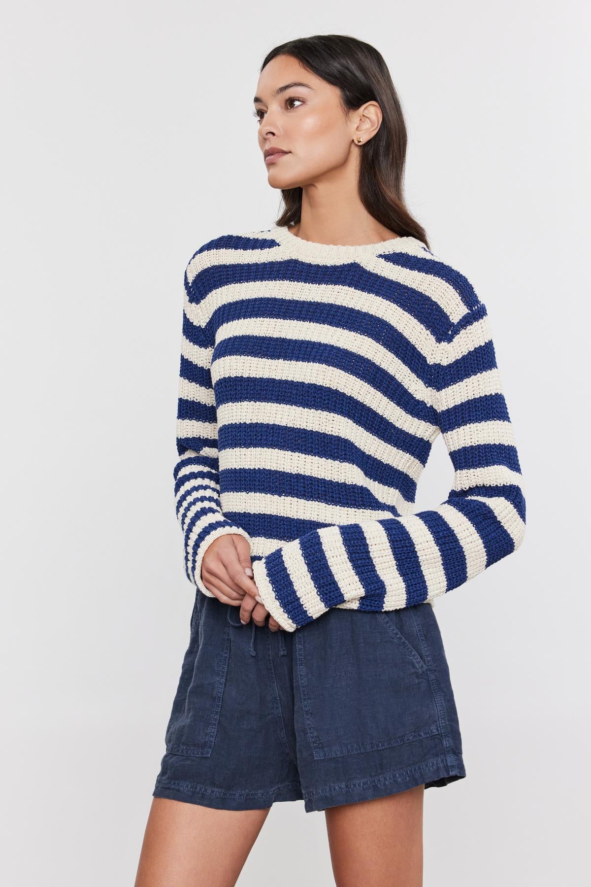   A woman in a Velvet by Graham & Spencer MAXINE SWEATER and denim skirt poses with her hand on her hip, looking to the side on a white background. 