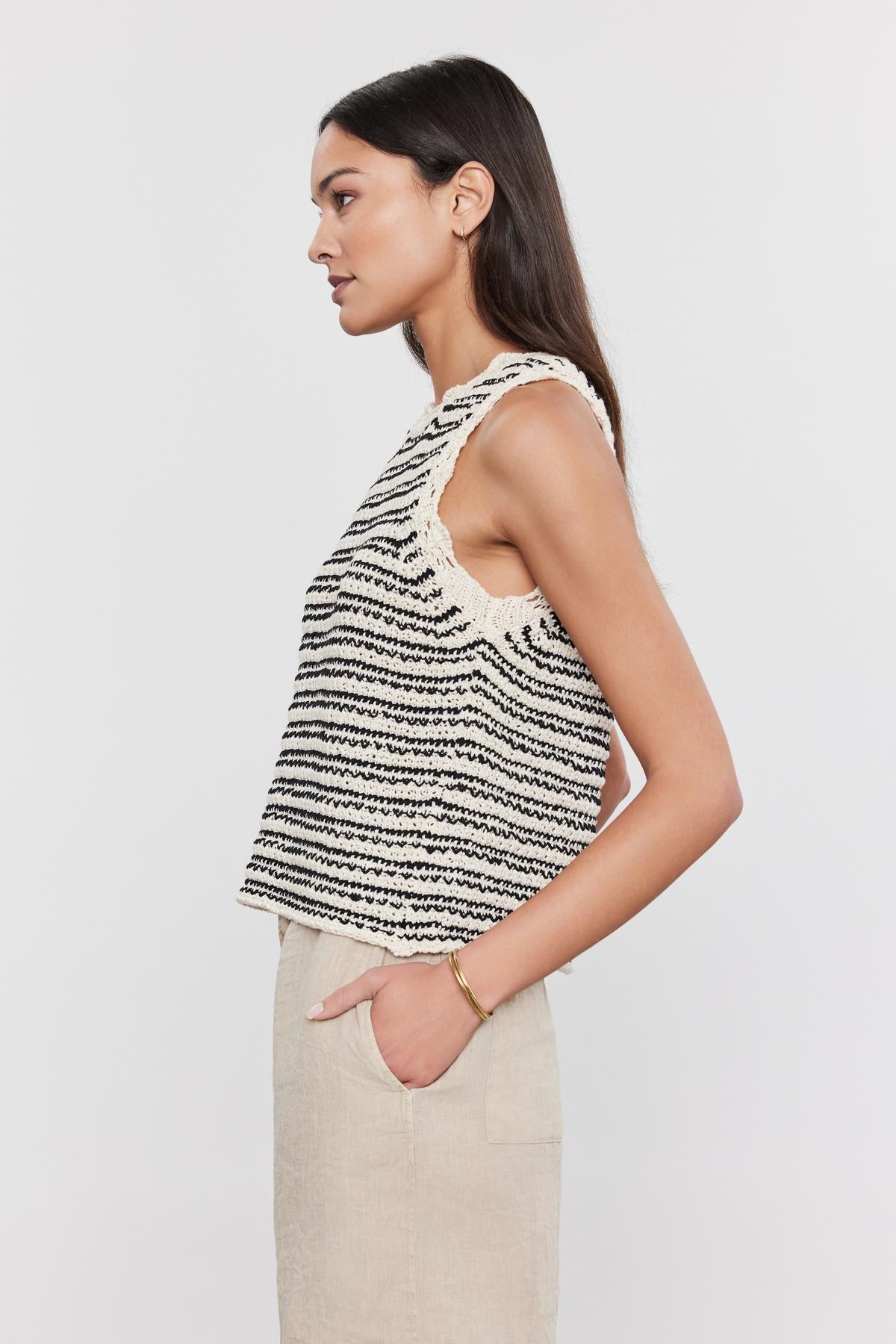  Side profile of a woman in a sleeveless, striped SOPHIE SWEATER VEST with scallop details and beige trousers against a white background by Velvet by Graham & Spencer. 