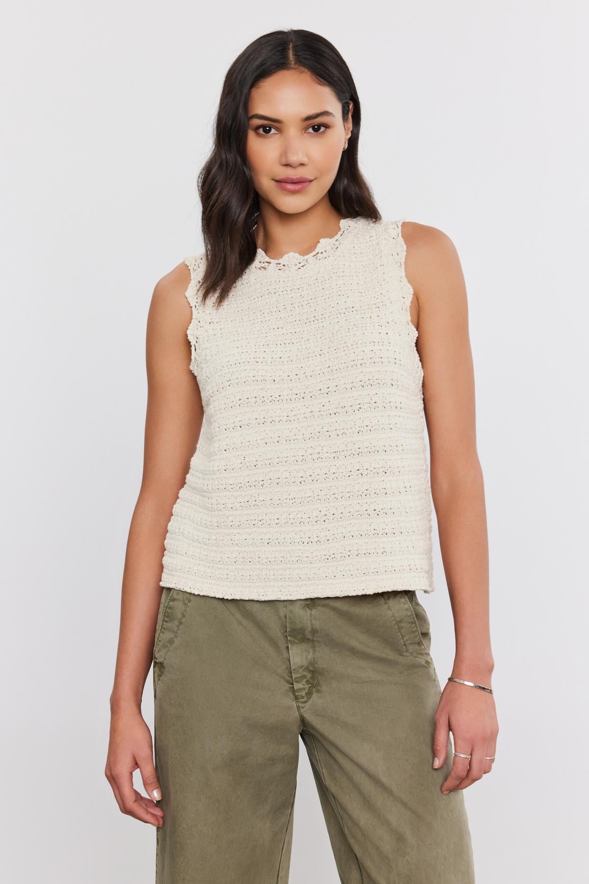   A person stands against a plain background wearing a sleeveless, cream-colored Velvet by Graham & Spencer SOPHIE SWEATER VEST with scallop details and green pants. 
