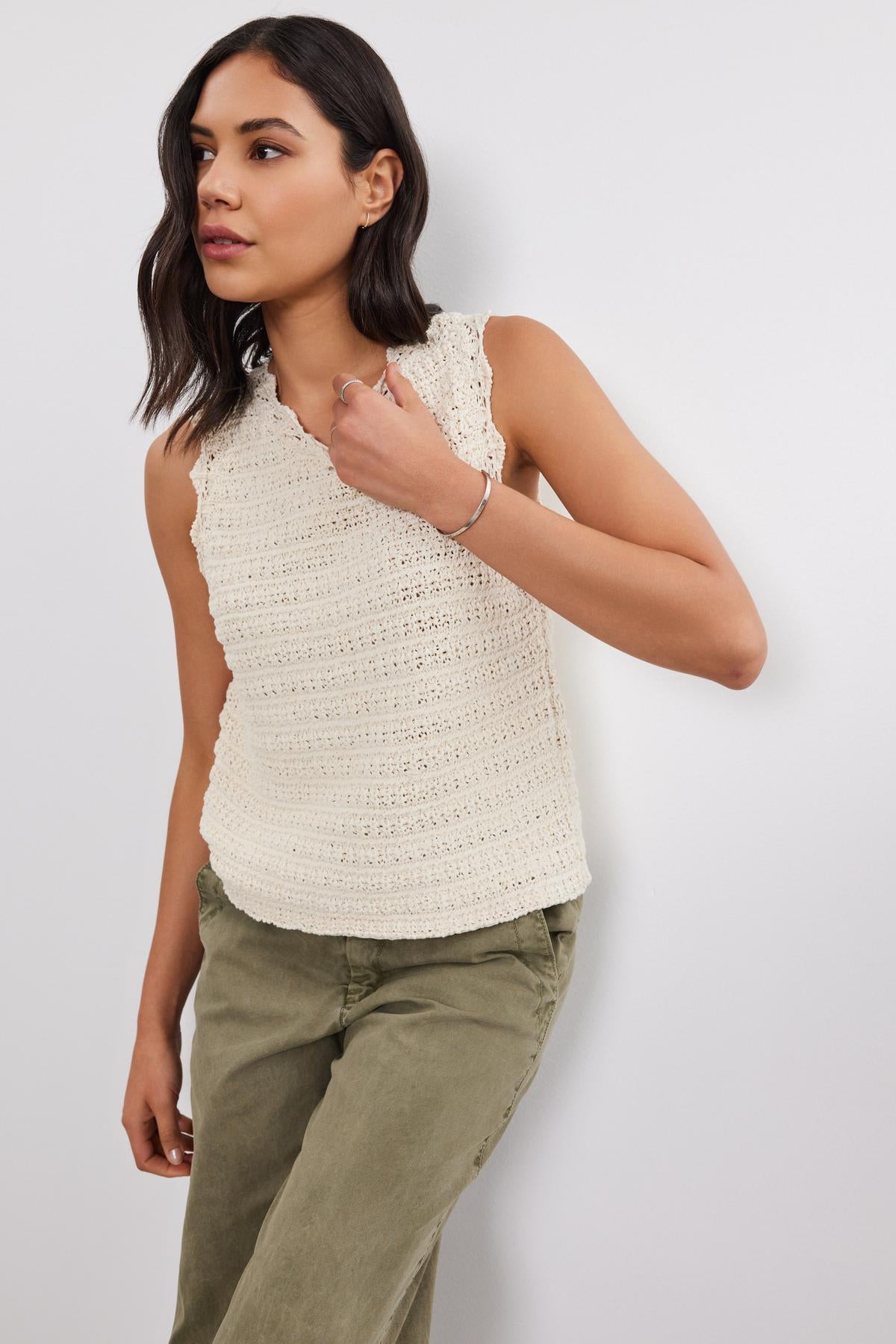 A person wearing a sleeveless, cream-colored SOPHIE SWEATER VEST by Velvet by Graham & Spencer with scallop details and olive green pants stands against a white wall.-37054560370881