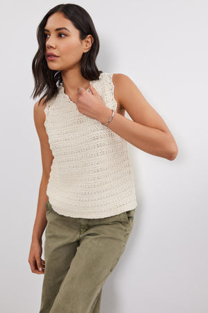 A person wearing a sleeveless, cream-colored SOPHIE SWEATER VEST by Velvet by Graham & Spencer with scallop details and olive green pants stands against a white wall.