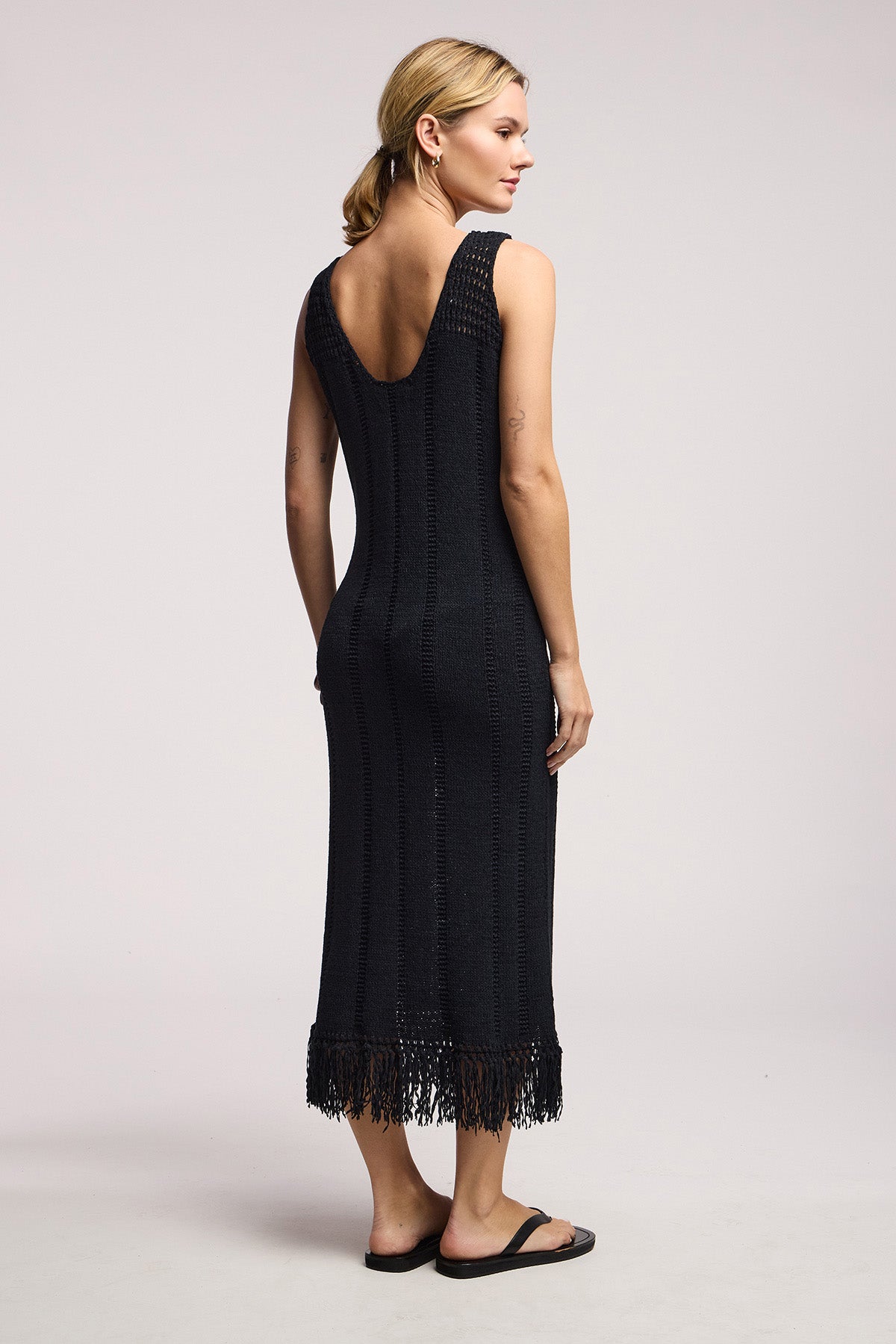 A woman, facing away, wears Velvet by Graham & Spencer’s sleeveless black TOMA DRESS—a crochet maxi with low back and fringe hem—paired with black sandals against a plain light background.-41527525343425