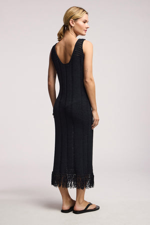 A woman, facing away, wears Velvet by Graham & Spencer’s sleeveless black TOMA DRESS—a crochet maxi with low back and fringe hem—paired with black sandals against a plain light background.