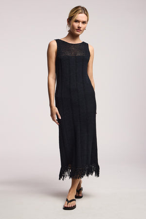 A woman wears the TOMA DRESS by Velvet by Graham & Spencer—a sleeveless black crochet summer dress with a fringed hem—paired with black sandals, standing against a plain background.