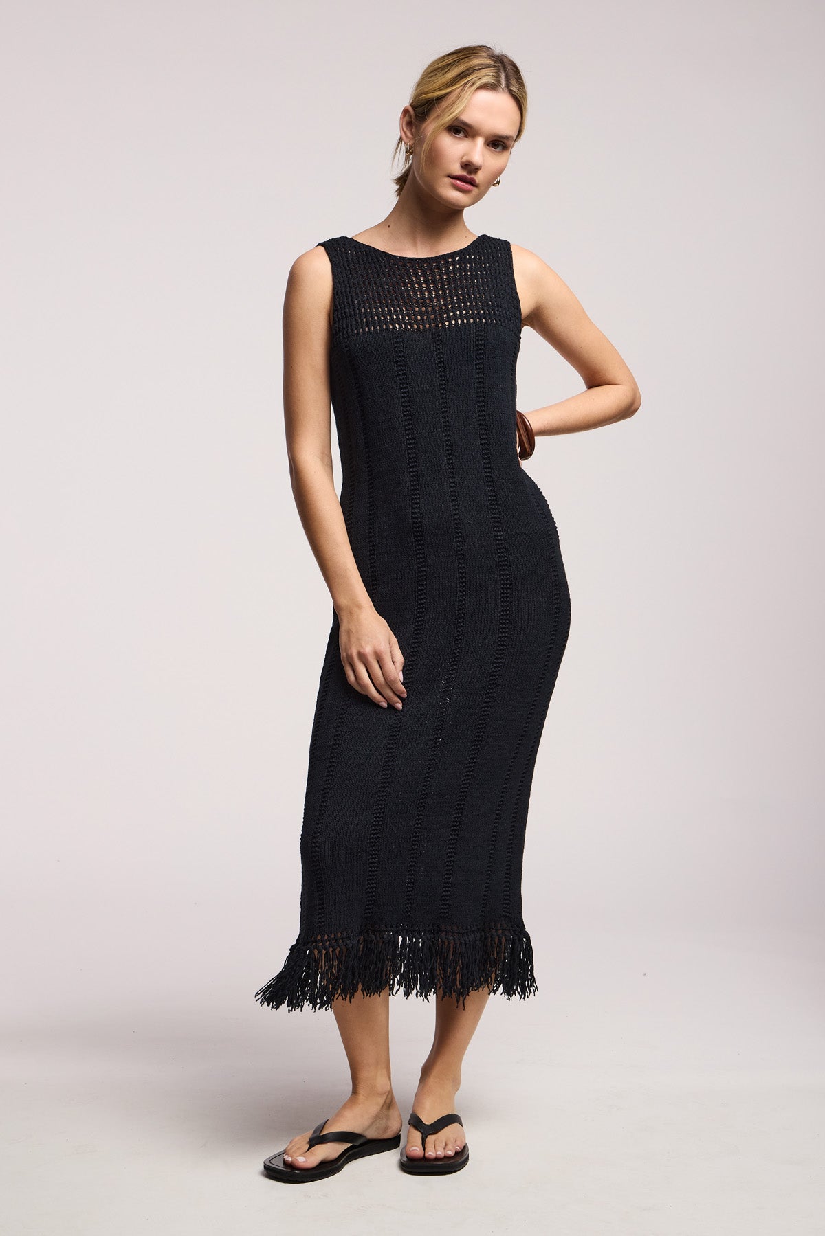 A woman models the Velvet by Graham & Spencer TOMA DRESS, a sleeveless black crochet design with fringe at the hem, paired with black sandals against a plain light background.-41527525114049