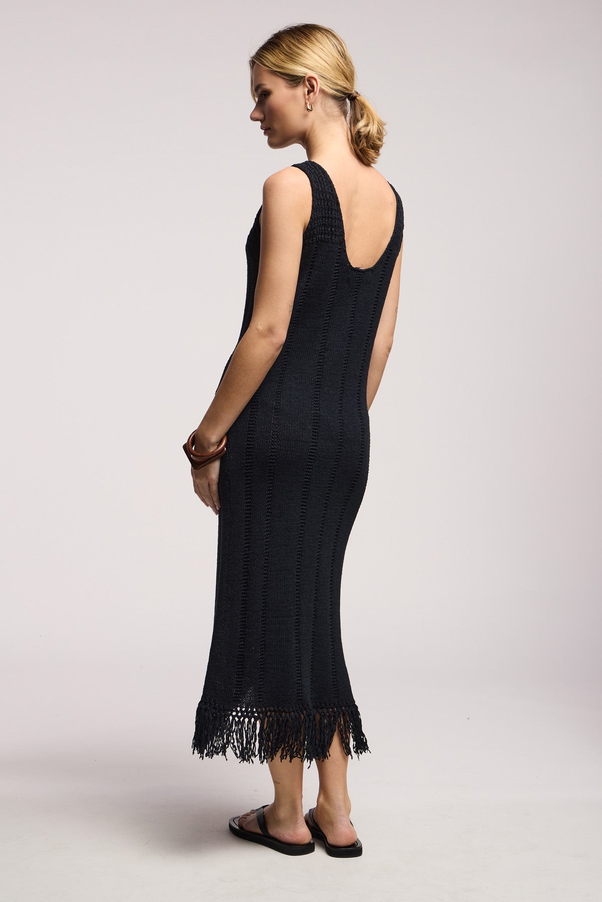 A woman stands sideways against a plain light background wearing the TOMA DRESS by Velvet by Graham & Spencer—a black sleeveless crochet dress with a low back and fringe hem—paired with black sandals and simple jewelry.