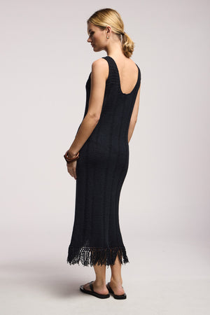 A woman stands sideways against a plain light background wearing the TOMA DRESS by Velvet by Graham & Spencer—a black sleeveless crochet dress with a low back and fringe hem—paired with black sandals and simple jewelry.