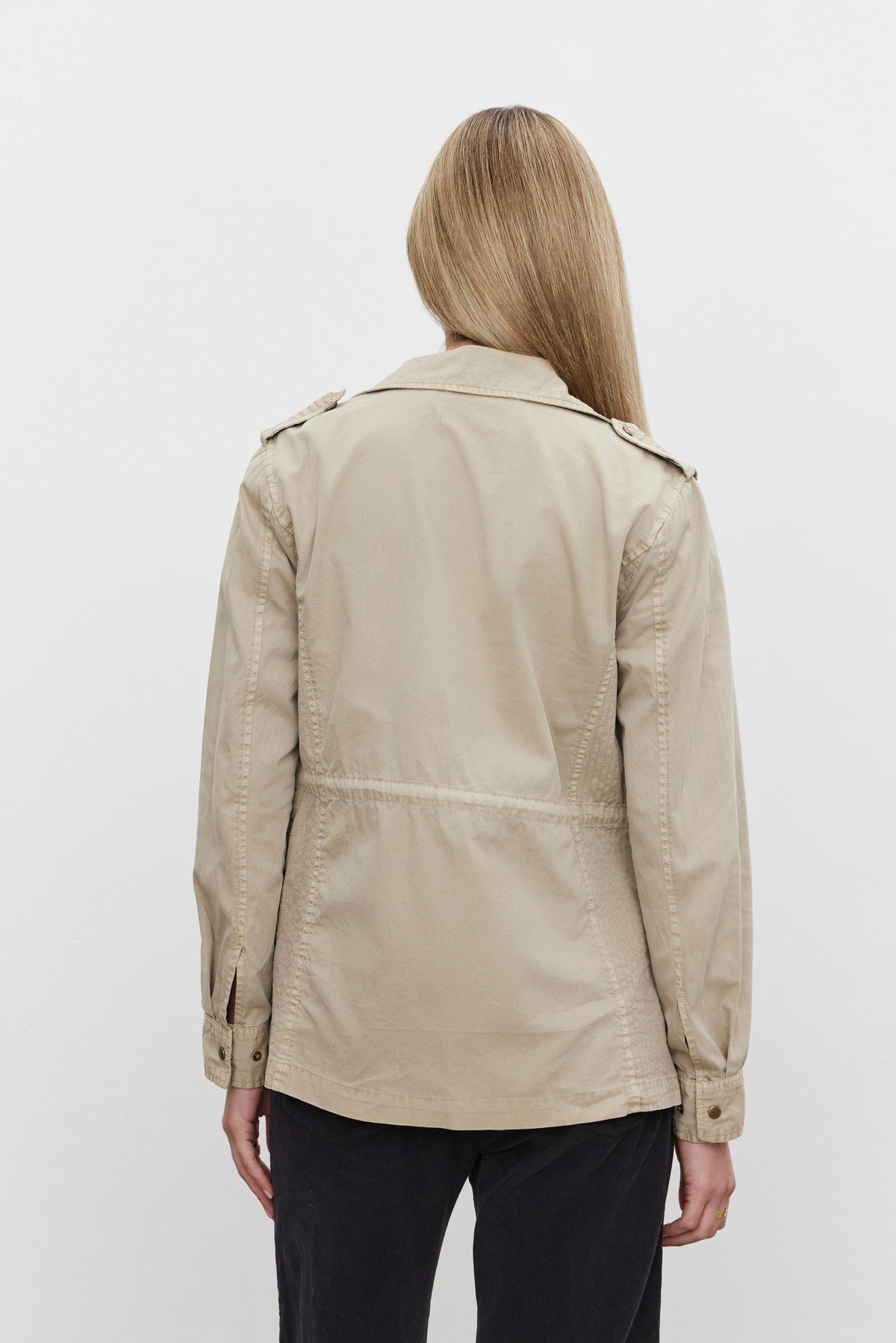   A person with long hair is wearing the RUBY LIGHT-WEIGHT ARMY JACKET from Velvet by Graham & Spencer in a beige cotton twill, paired with black pants, and is facing away from the camera against a plain background. 