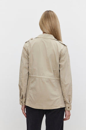 A person with long hair is wearing the RUBY LIGHT-WEIGHT ARMY JACKET from Velvet by Graham & Spencer in a beige cotton twill, paired with black pants, and is facing away from the camera against a plain background.