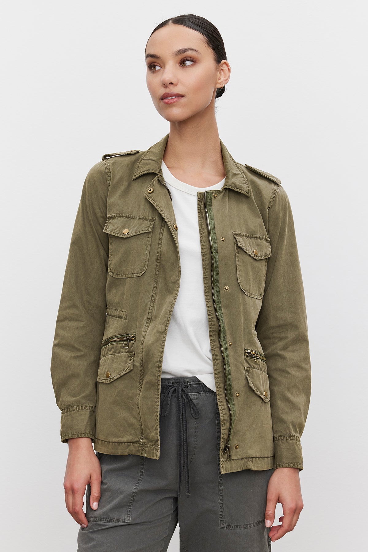   A woman embodies off-duty glamour as she stands against a plain white background, wearing the Velvet by Graham & Spencer RUBY LIGHT-WEIGHT ARMY JACKET in olive green over a white shirt and grey pants. 