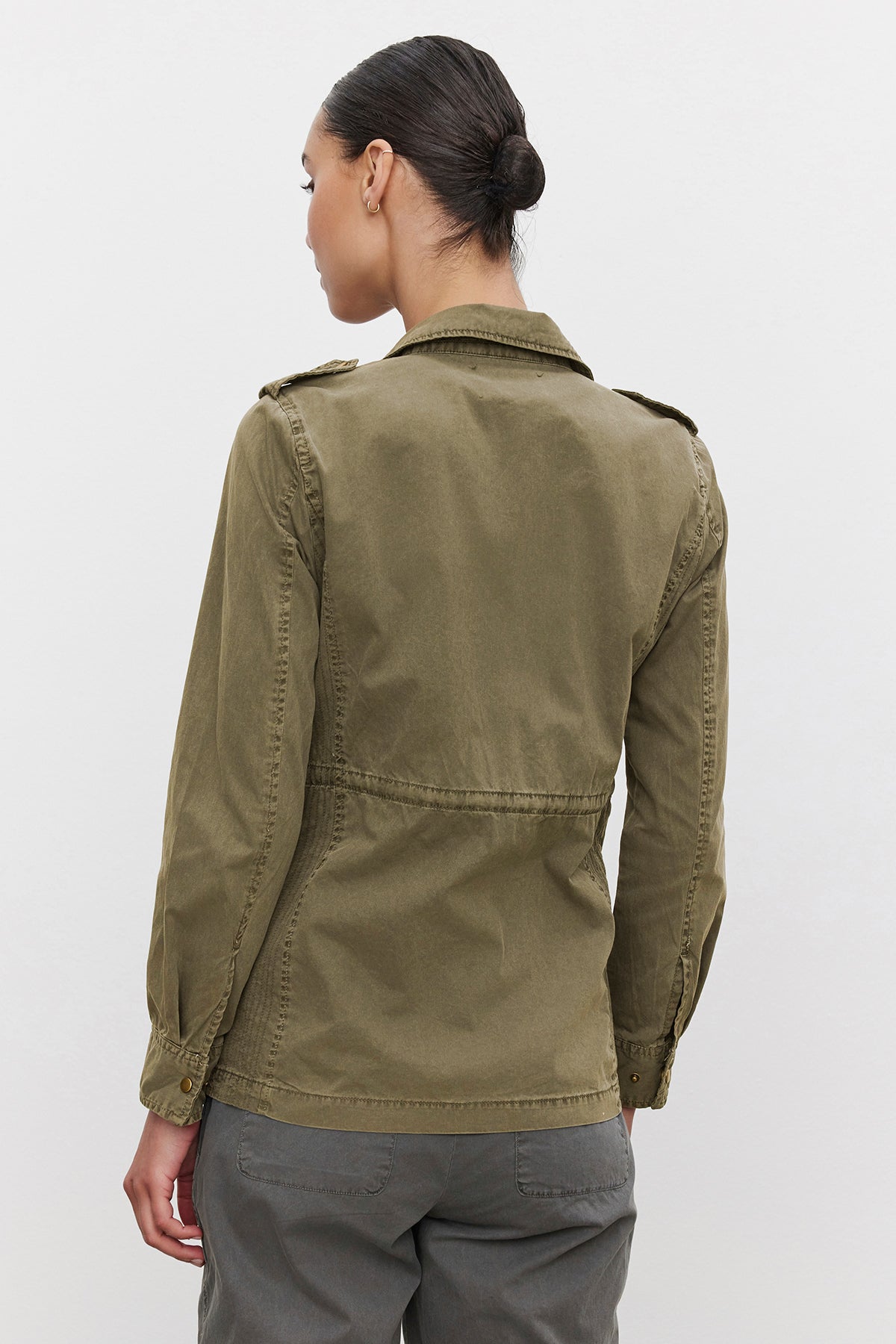   A person with hair tied back is standing on a plain background, viewed from behind, wearing the Velvet by Graham & Spencer RUBY LIGHT-WEIGHT ARMY JACKET in olive green and gray pants, exuding off-duty glamour. 