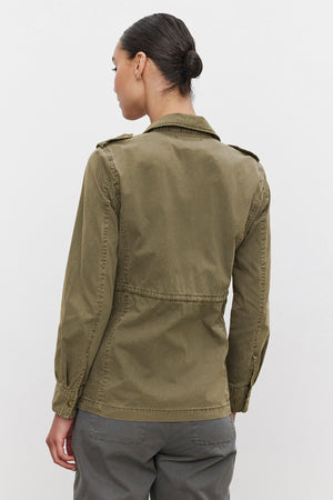 A person with hair tied back is standing on a plain background, viewed from behind, wearing the Velvet by Graham & Spencer RUBY LIGHT-WEIGHT ARMY JACKET in olive green and gray pants, exuding off-duty glamour.