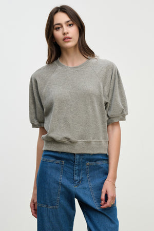 A woman faces the camera against a plain light background, wearing the AVIS PULLOVER by Velvet by Graham & Spencer—a grey cotton velour crewneck—paired with blue jeans.