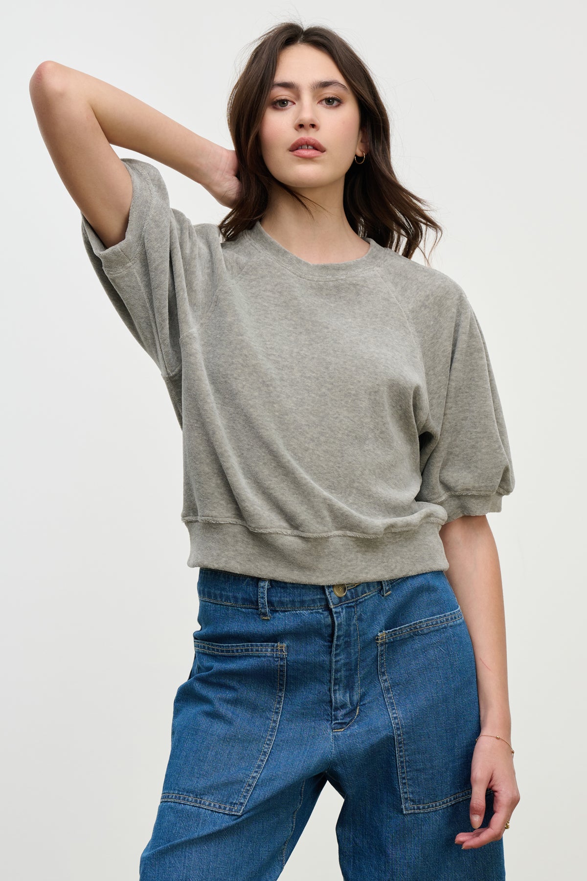A woman stands against a plain light background with one arm bent behind her head, wearing the AVIS PULLOVER by Velvet by Graham & Spencer—a loose grey cotton velour crewneck—paired with blue jeans.-39814820724929