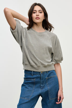 A woman stands against a plain light background with one arm bent behind her head, wearing the AVIS PULLOVER by Velvet by Graham & Spencer—a loose grey cotton velour crewneck—paired with blue jeans.