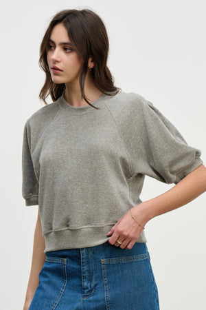 A woman in the AVIS PULLOVER by Velvet by Graham & Spencer— a loose grey cotton velour top with raglan puff sleeves— pairs it with a blue denim skirt, standing against a plain light background.