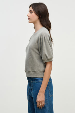 A woman stands in profile against a plain background, wearing Velvet by Graham & Spencer's AVIS PULLOVER in light gray cotton velour with raglan puff sleeves, paired with blue jeans.
