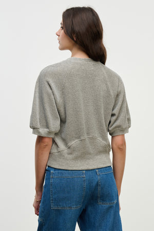 A woman with long brown hair stands with her back to the camera against a plain light background, wearing the AVIS PULLOVER by Velvet by Graham & Spencer in gray cotton velour with a crew neckline and blue jeans.