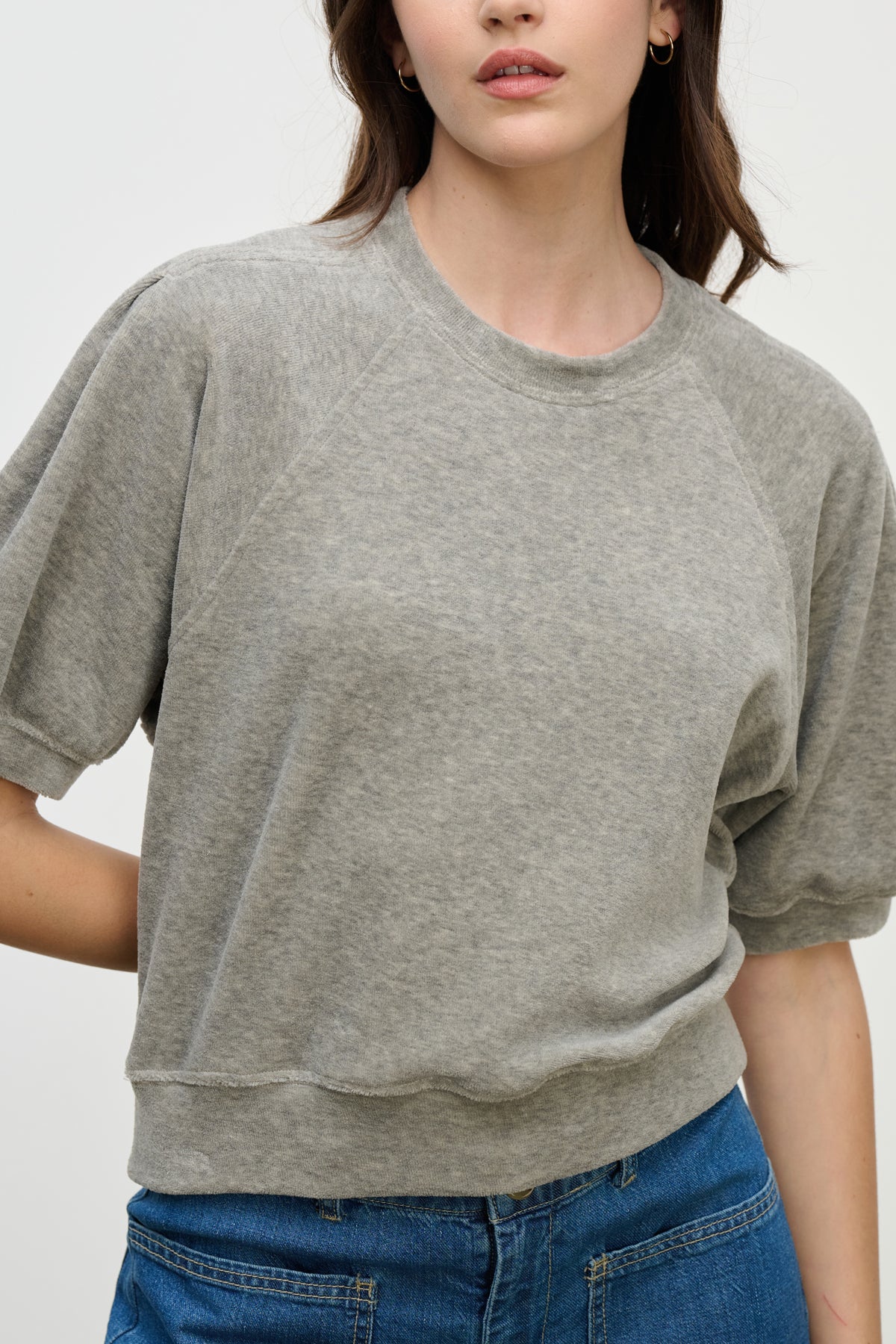 A person wearing the AVIS PULLOVER by Velvet by Graham & Spencer, a light grey cotton velour crew-neck top, pairs it with blue jeans while standing against a plain background.-39814820593857