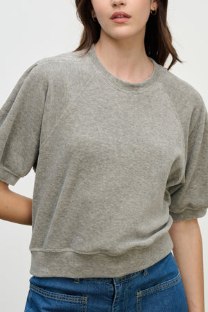A person wearing the AVIS PULLOVER by Velvet by Graham & Spencer, a light grey cotton velour crew-neck top, pairs it with blue jeans while standing against a plain background.